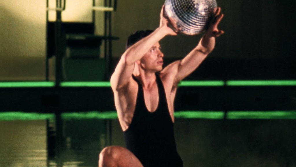 Zero (Normand Fauteux) balancing a disco ball on his arms next to a swimming pool. He is wearing a man's bathing suit.