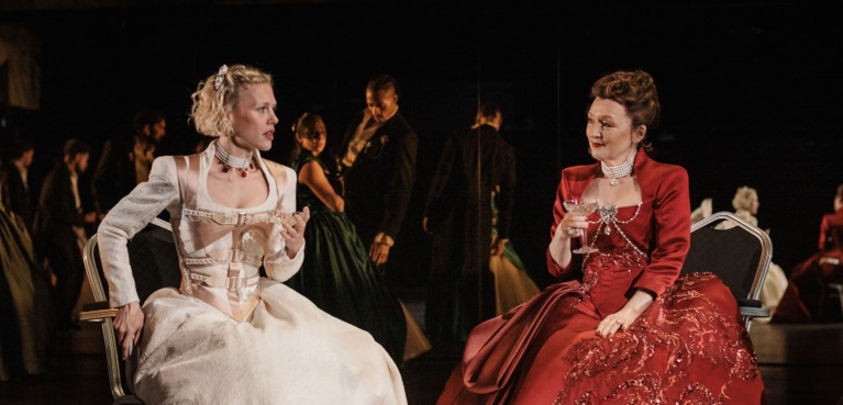 Cécile (Hannah van der Westhuysen) in a white dress, talking to the Marquise de Merteuil (Lesley Manville) who is wearing a red dress. Cécile looks somewhat scandalized. 