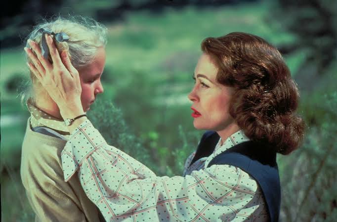 Christina (Mara Hobel) standing in front of a kneeling Joan (Faye Dunaway). Joan is petting her hair, Christina looks unhappy.