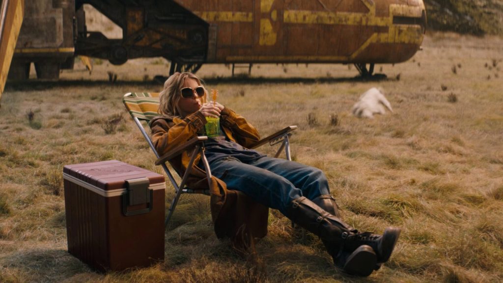 Kara aka Supergirl (Milly Alcock) in a rickety lounge chair with a giant cocktail in hand. Behind her is Krypto, a white dog, flailing around in the grass.