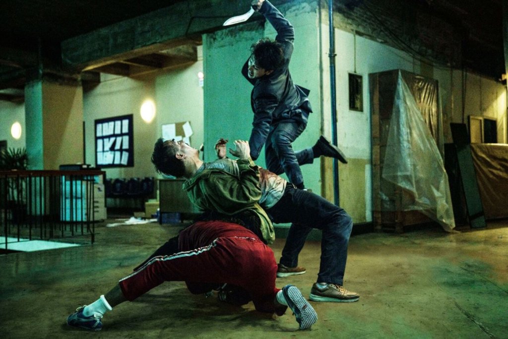 One person kneeling on the floor as another leans backwards over him and a third one is jumping in the air with a knife raised high, trying to stab the second person.