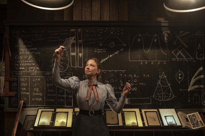Katharine (Haley Bennett) in front of a blackboard covered in numbers and diagrams. She is holding up a glass plate to the light.