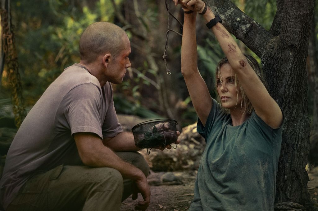 Ben (Taron Egerton) crouching in front of Sasha (Charlize Theron) who is tied up against a tree with her hands above her head. He is holding a basket with something, probably food. They are making intense, hostile eye-contact.