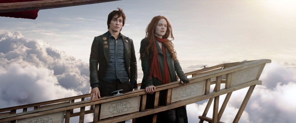 Tom (Robert Sheehan) and Hester (Hera Hilmar) standing on a narrow wooden platform that is part of a flying airship.