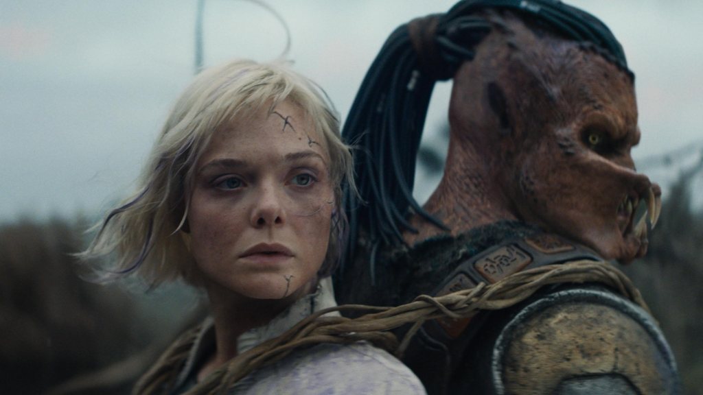 Dek (Dimitrius Schuster-Koloamatangi) back to back with Thia (Elle Fanning)- She is fastened to his back with crude ropes.