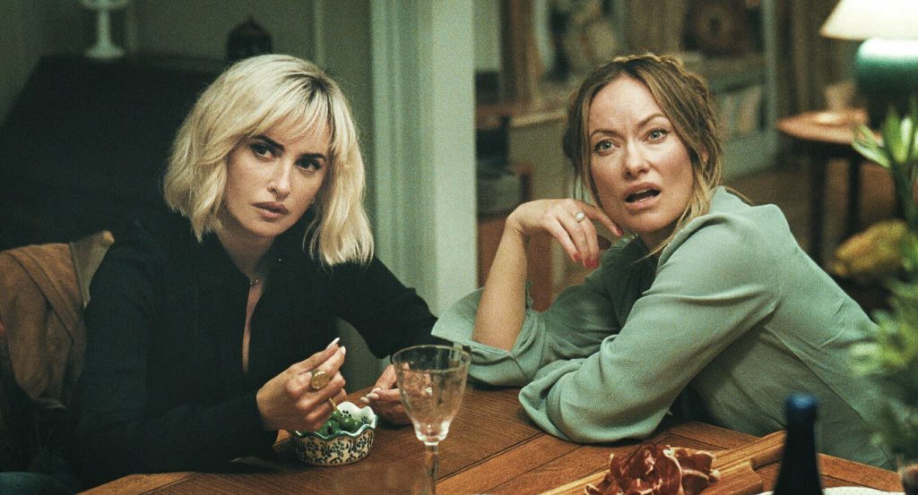 Piña (Penélope Cruz) and Angela (Olivia Wilde) sitting close together at a table. Both are looking up with varying shades of critical expressions.