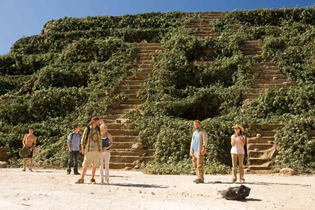 The group of young people standing in front of a pyramid that is covered in vines apart from two paths. They are all turned away from the structure, though.